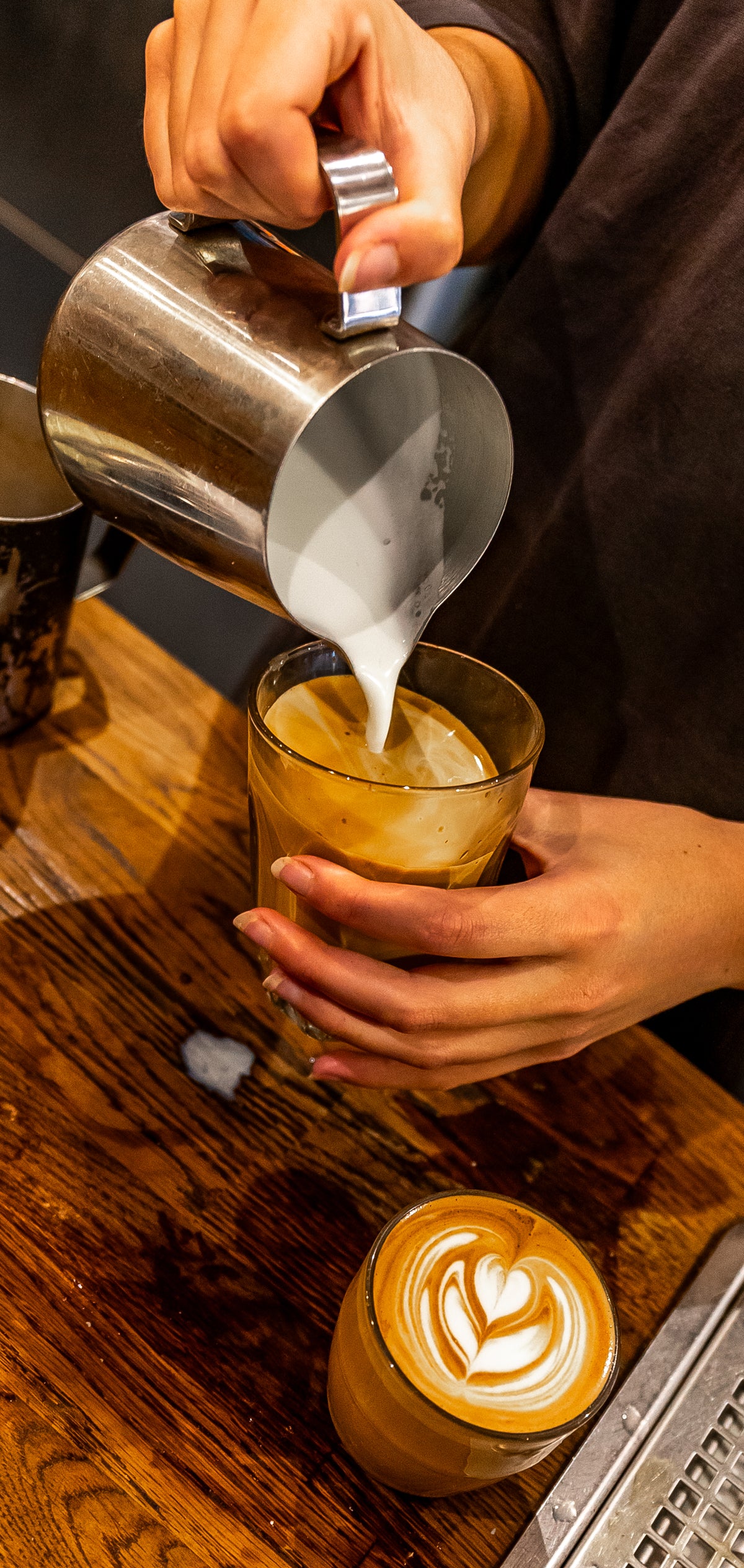 A close-up of a barista pouring steamed milk from a stainless steel pitcher into a glass of Speciality coffee on a wooden counter in Brighton, with another latte featuring heart-shaped latte art in the foreground. 